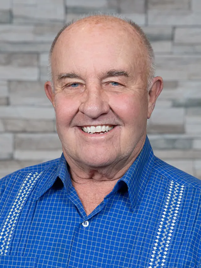 Smiling elderly man wearing a blue checkered shirt with a stone wall background.