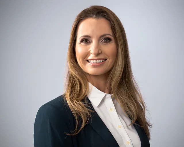 Professional woman with long hair wearing a dark blazer and white shirt smiling against a gray background