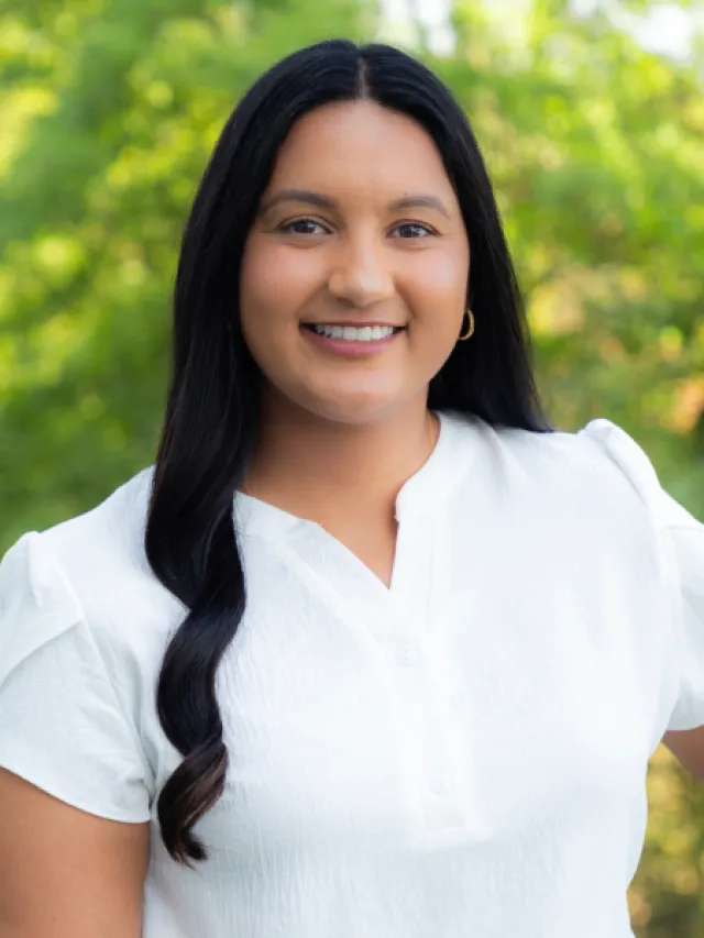 Smiling young woman with long black hair wearing a white blouse standing outdoors with green foliage background