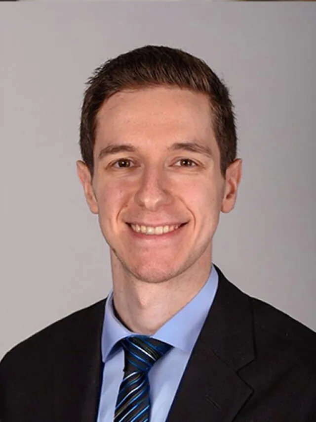 Professional headshot of a young man smiling, wearing a black suit, blue shirt, and striped tie against a gray background.