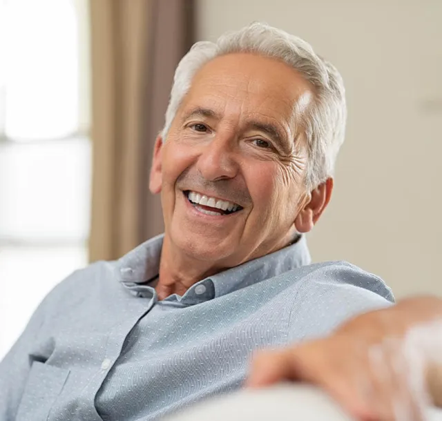 Smiling elderly man with gray hair wearing a light blue shirt, relaxing indoors on a beige sofa.