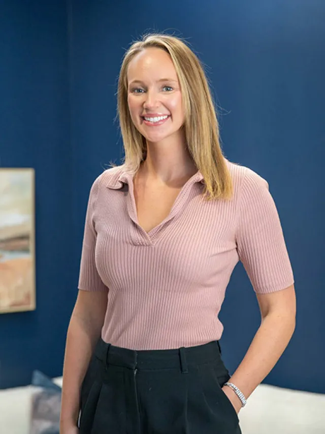 Smiling woman with blonde hair wearing a pink ribbed shirt and black pants standing against a blue wall.