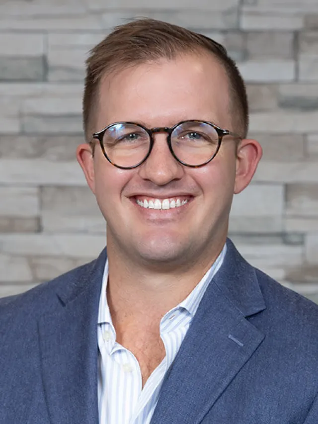 Young man with glasses smiling, wearing a blue suit jacket and white striped shirt against a stone wall background.