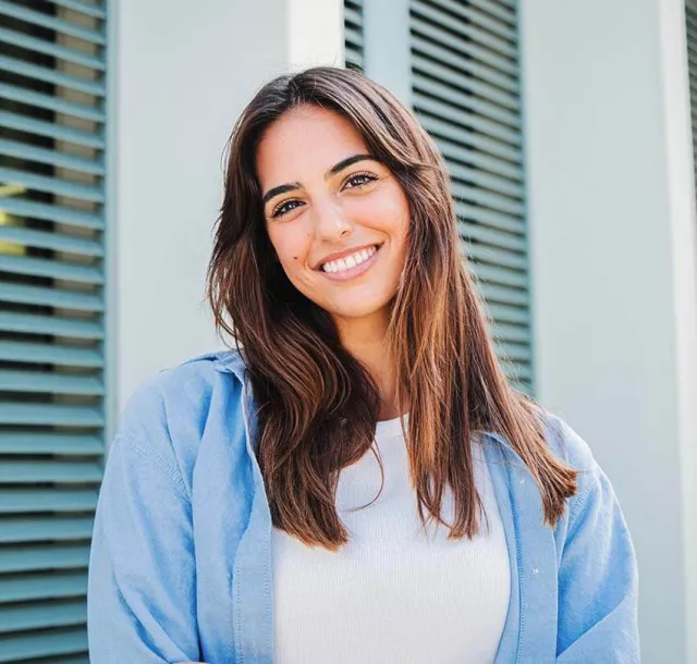 Smiling young woman with long hair wearing a blue shirt, standing in front of green shutters.