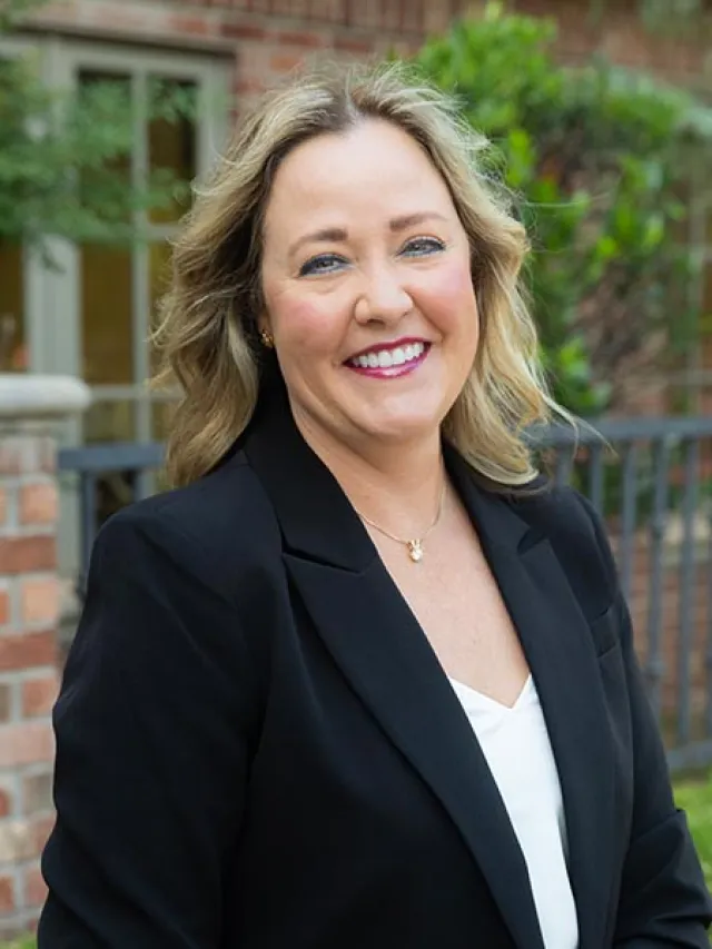 Smiling woman with blonde hair wearing a black blazer and white top outdoors by a brick building and greenery.