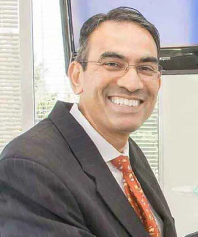 Smiling man in business attire with glasses against an office background.
