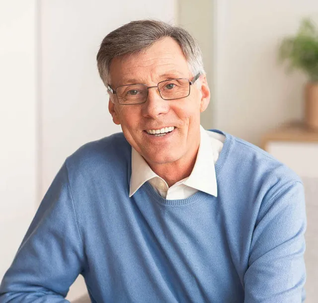 Smiling elderly man wearing glasses and a blue sweater sitting indoors with a bright background.