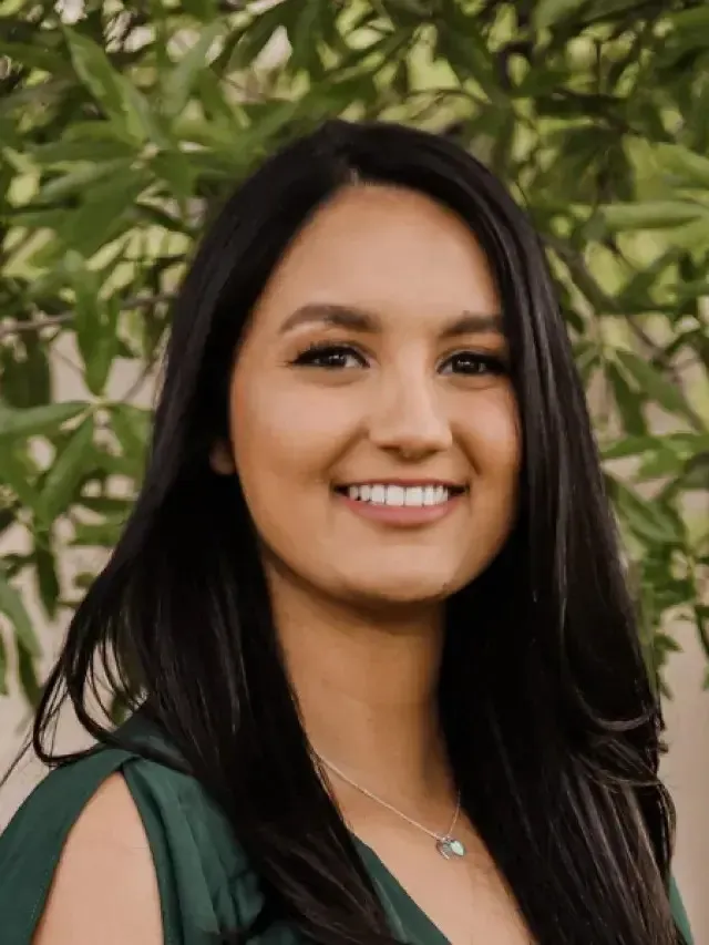 Smiling young woman with long black hair and green top standing outdoors with leafy background