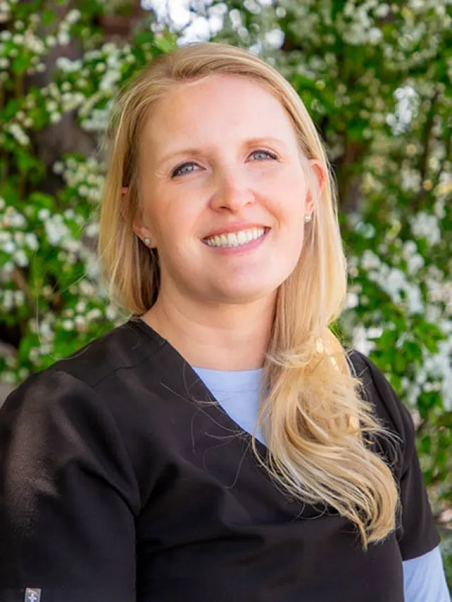 Smiling woman with blonde hair wearing black scrubs standing outdoors with greenery and white flowers in background