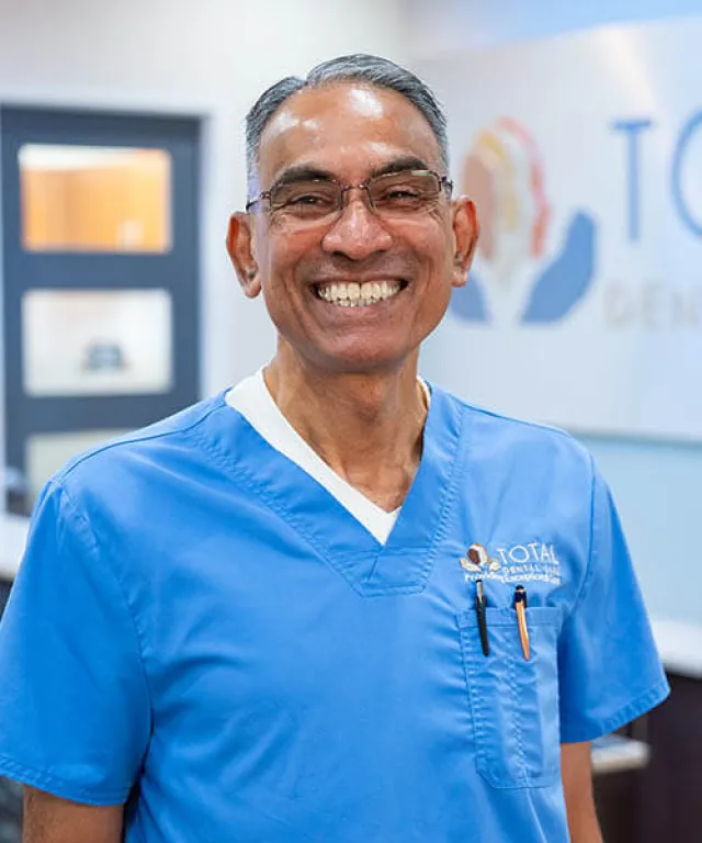 Smiling male dentist in blue scrubs and glasses standing in dental clinic with Total Dental logo in background.