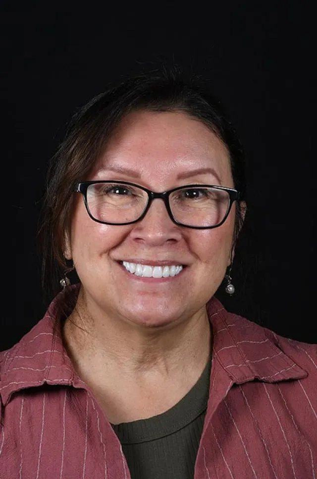 Smiling middle-aged woman with glasses wearing a red shirt and pearl earrings against a black background