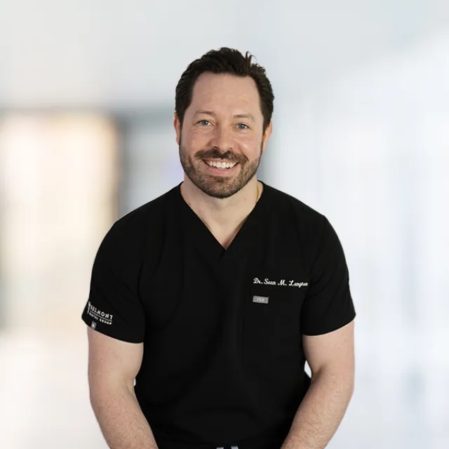 Smiling male healthcare professional in black scrubs standing in a bright, modern medical office.