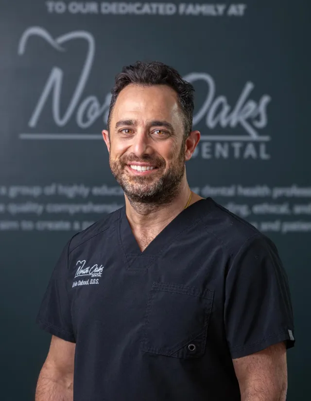 Smiling male dentist in black scrubs standing in front of North Oaks Dental sign with beard and short hair.