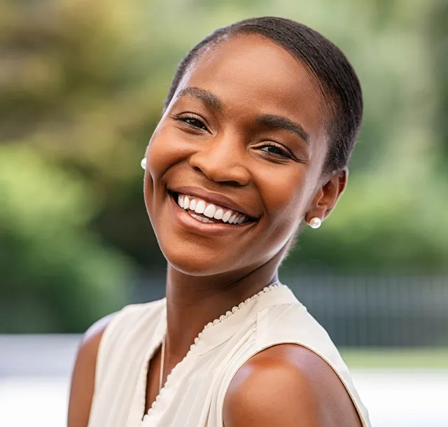 Smiling young woman with pearls and white sleeveless top in outdoor setting with blurred green background