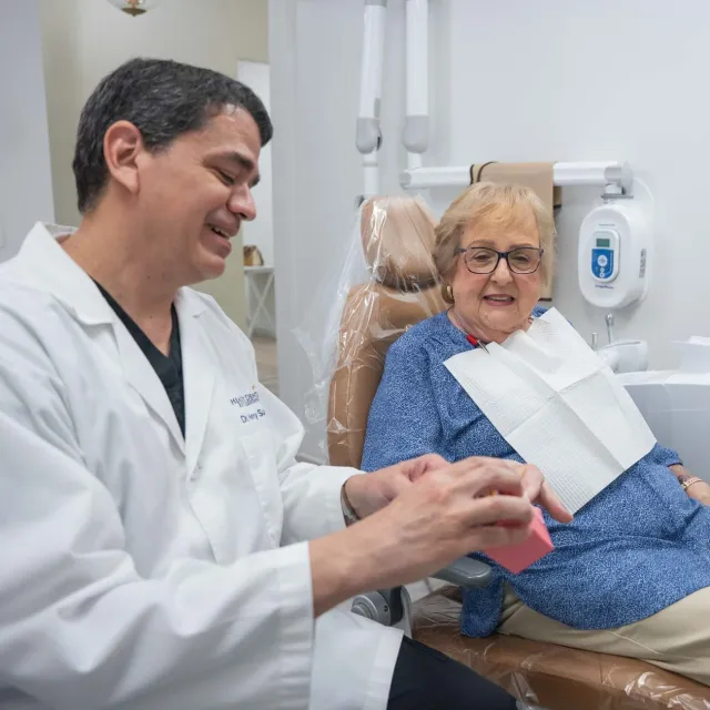 Smiling elderly man with gray hair wearing a light blue shirt, relaxing indoors on a beige sofa.