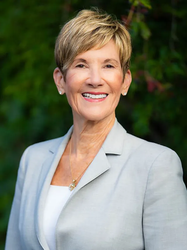 Smiling mature woman with short blonde hair wearing a light gray blazer and necklace outdoors