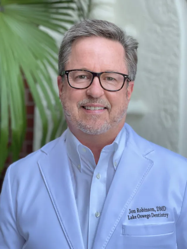 Middle-aged man with glasses wearing a white dental coat standing in front of greenery and white wall.