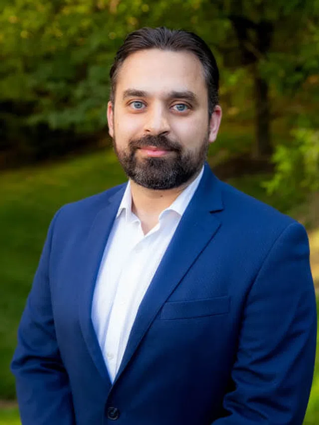 Smiling man with beard wearing a blue suit and white shirt outdoors with green trees in the background