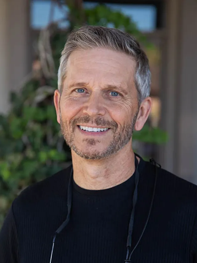 Smiling middle-aged man with gray hair and beard wearing a black shirt, standing outdoors with green plants background