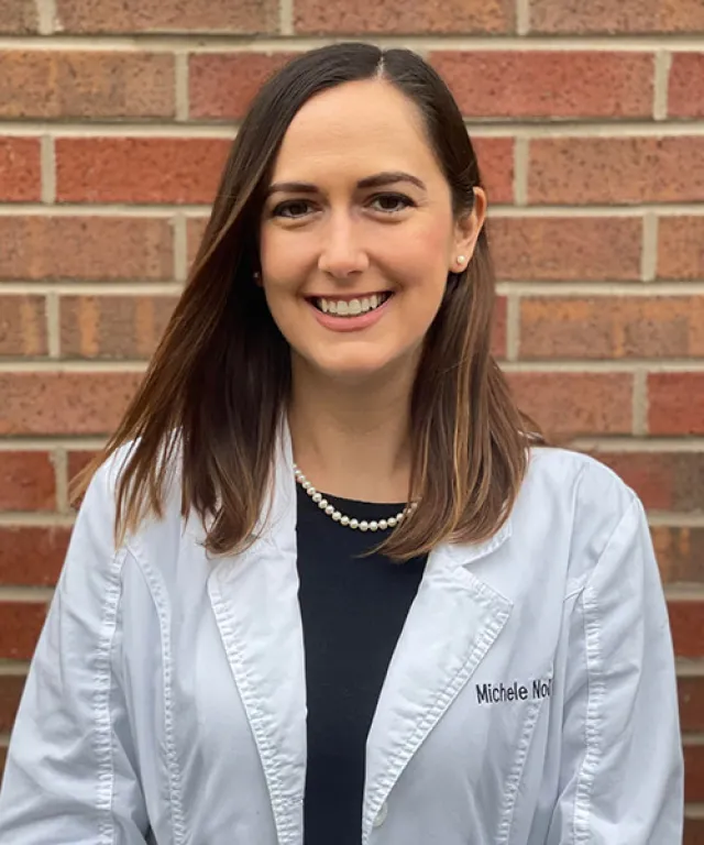 Smiling woman with long brown hair wearing a white lab coat and pearl necklace against brick wall.