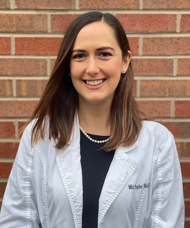 Smiling woman in white medical coat and pearl necklace standing against a brick wall background