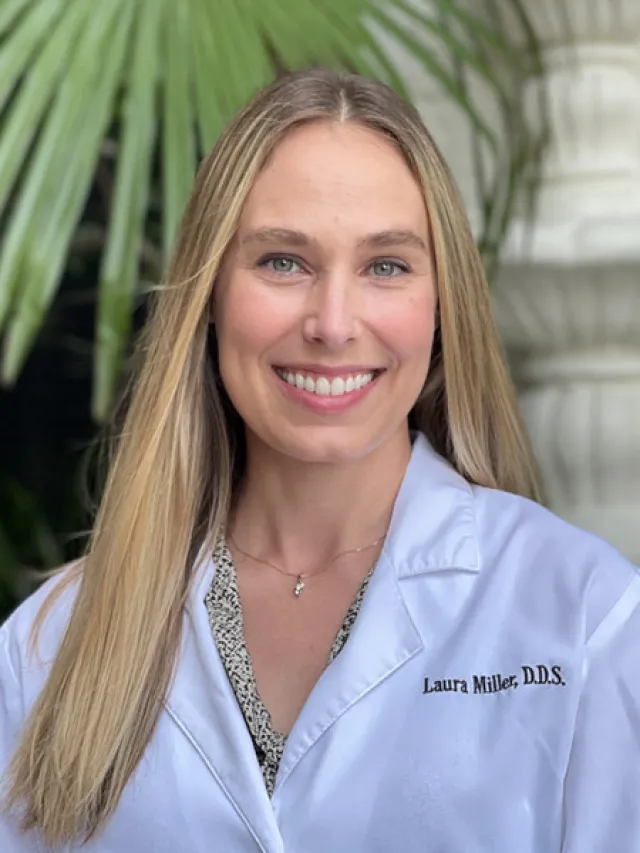 Smiling female dentist with long blonde hair in a white coat beside green plants and stone background