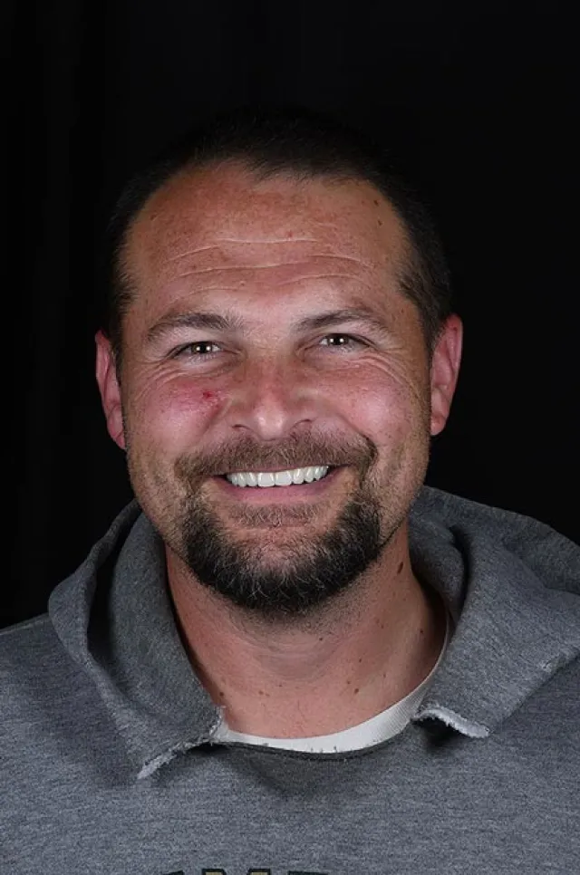 Smiling man with short dark hair, beard, and mustache wearing gray hoodie against a black background