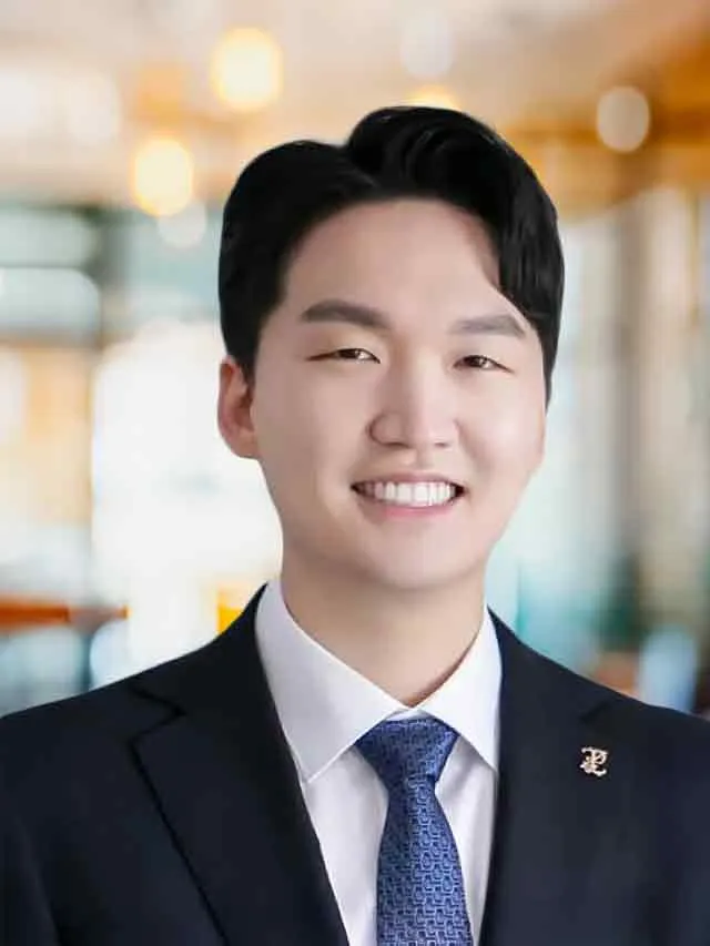 Young man in formal suit and blue tie smiling with blurred indoor background of warm lighting.