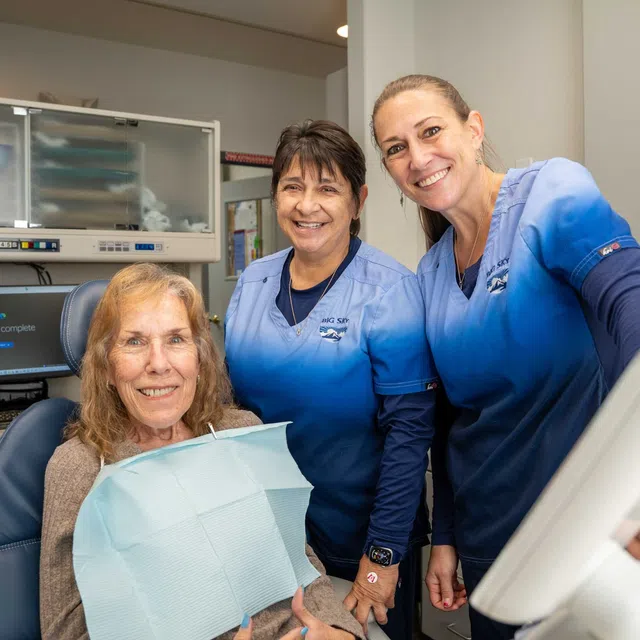Smiling dental patient with two female dental professionals in blue scrubs in the clinic.