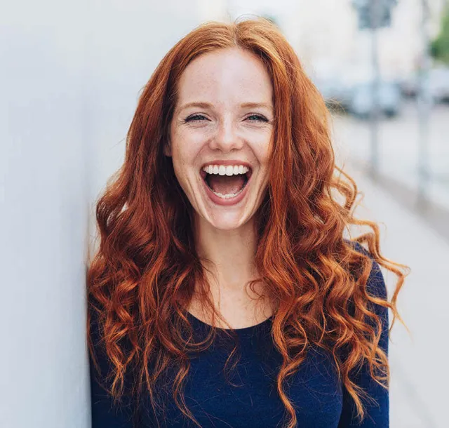 Joyful red-haired woman with curly hair laughing outdoors against a light background