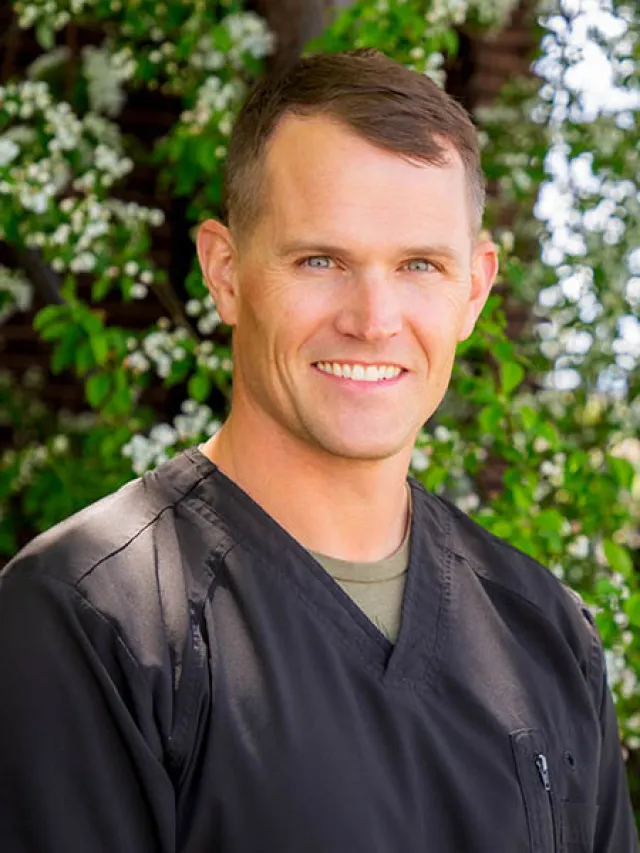 Smiling man in black medical scrubs standing outdoors with green foliage and white flowers in background