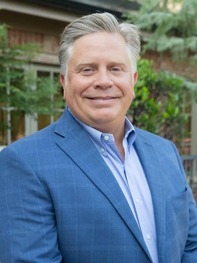Smiling middle-aged man in a blue suit jacket and light blue shirt standing outdoors with greenery in the background