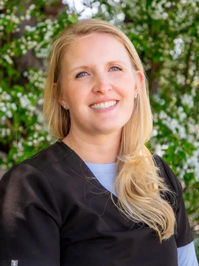 Smiling blonde woman in black scrubs standing outdoors with green foliage and white flowers in the background