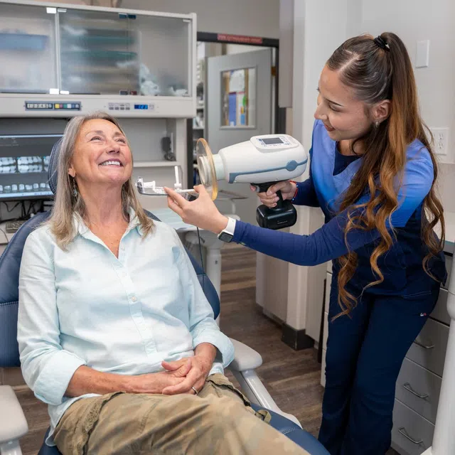 Dental professional uses handheld X-ray device on smiling seated patient in modern dental clinic.