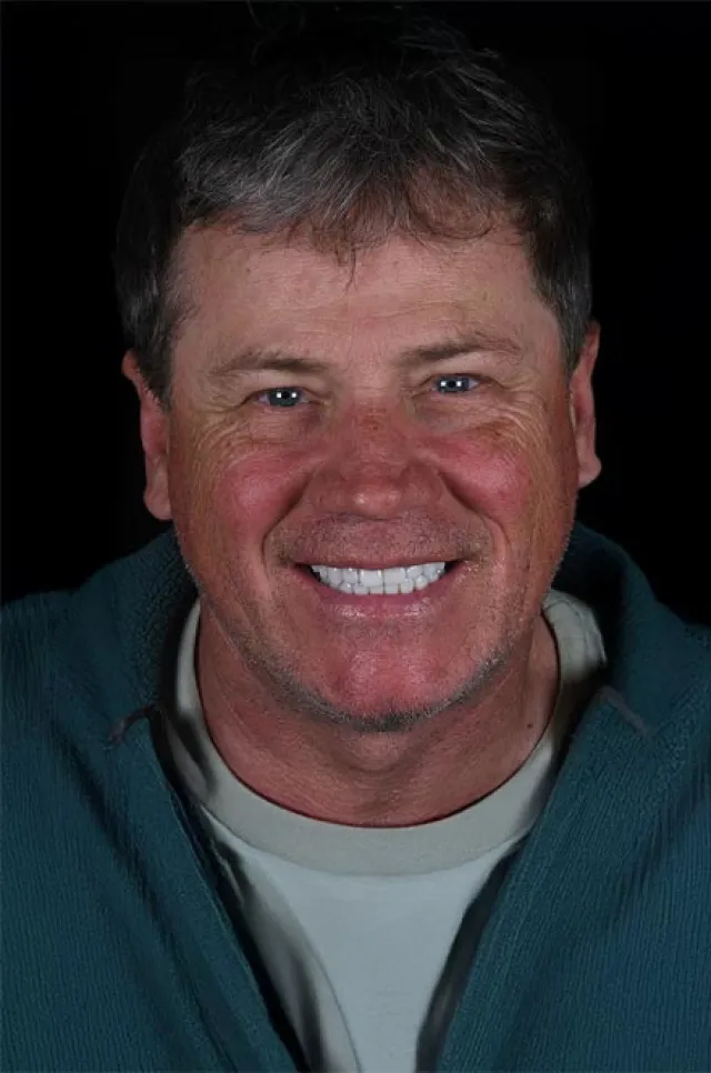 Middle-aged man with short graying hair smiles warmly, wearing a teal jacket over a beige shirt against a black background