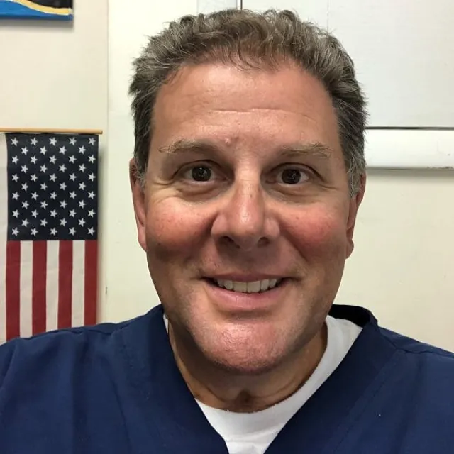 Smiling middle-aged man wearing navy scrubs with an American flag in the background indoors.