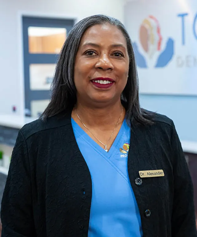 Smiling female doctor wearing blue scrubs and black coat with a Dr. Alexander name tag in a medical office.