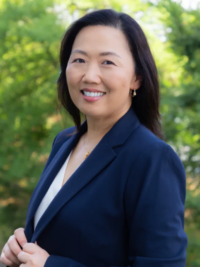 Smiling professional Asian woman in navy blazer posing outdoors with greenery background.