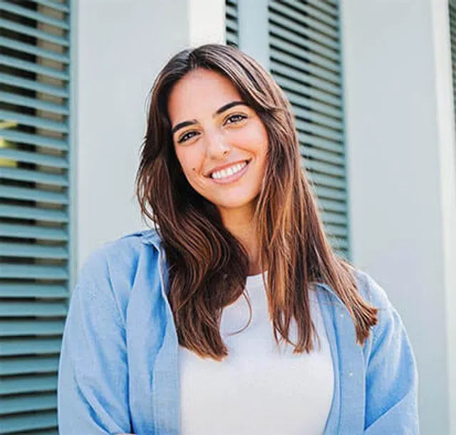 Smiling young woman with long hair wearing a blue shirt, standing in front of green shutters.