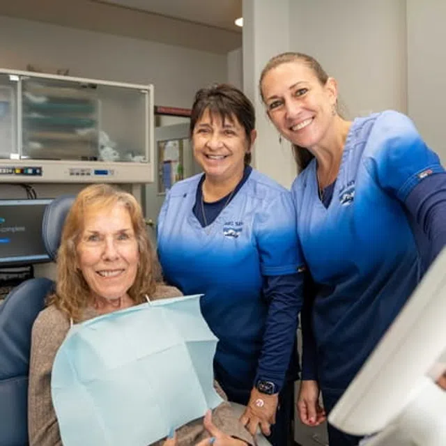 Smiling dental patient with two female dental professionals in blue scrubs in the clinic.