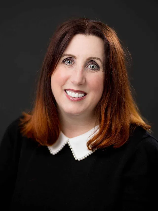 Smiling woman with red hair, pearl-adorned white collar, and black top against a dark background.