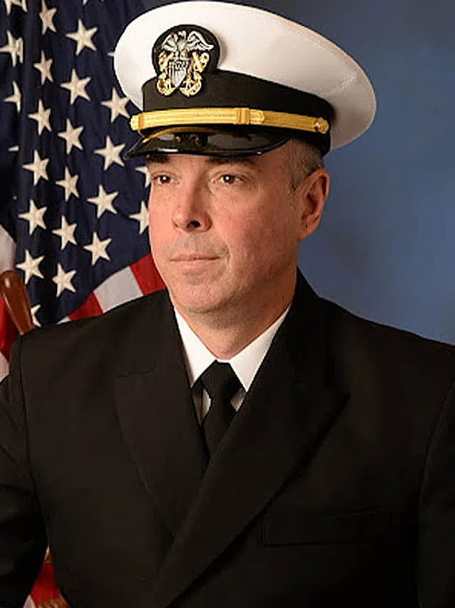 Portrait of a US Navy officer in formal uniform seated with folded hands, American flag and ship wheel in background