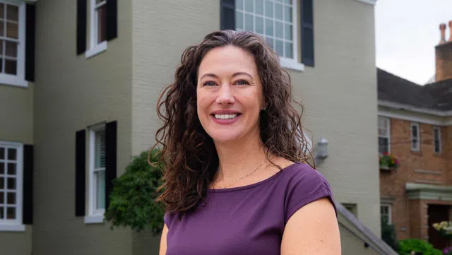Smiling woman with curly hair wearing a purple shirt standing outside a residential building on a clear day