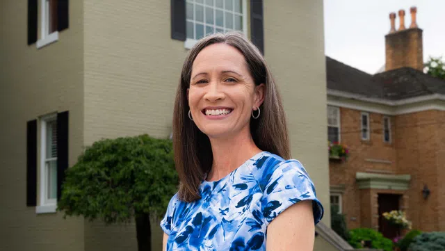 Smiling woman in blue floral dress stands outdoors in front of a residential house on a sunny day