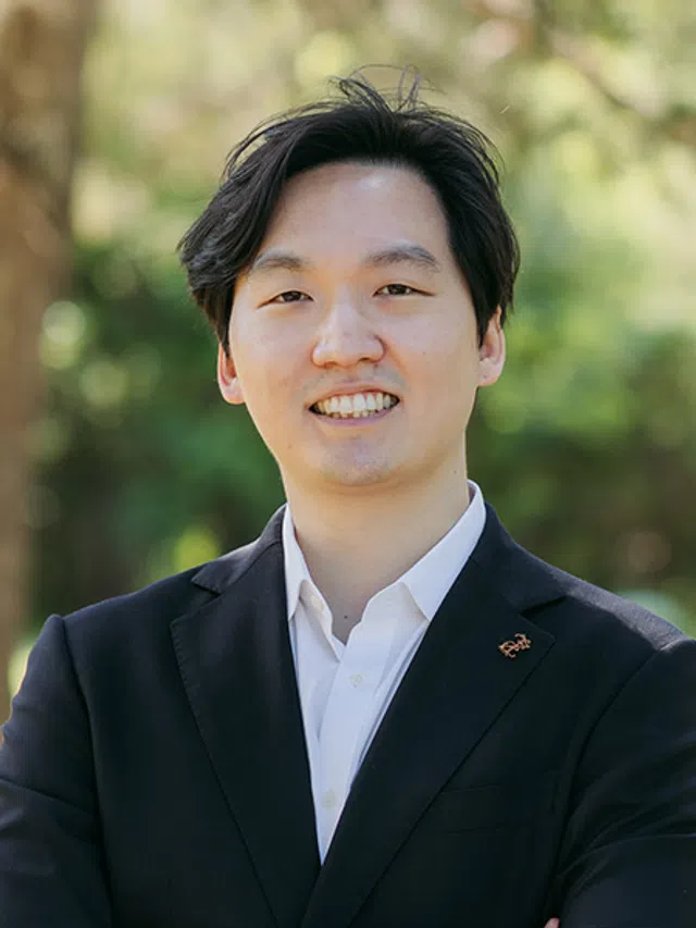 Smiling young man in a black suit and white shirt standing outdoors with blurred green background.