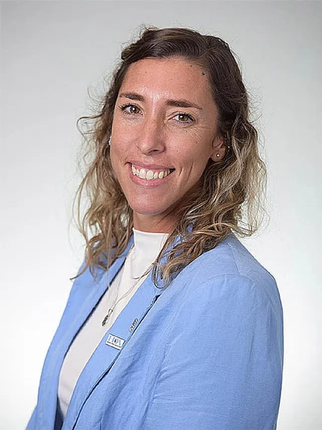 Smiling woman with curly hair wearing a light blue blazer and white top against a plain background