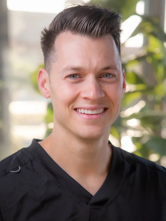 Smiling young man with blue eyes and styled dark hair wearing a black shirt in a bright indoor setting.