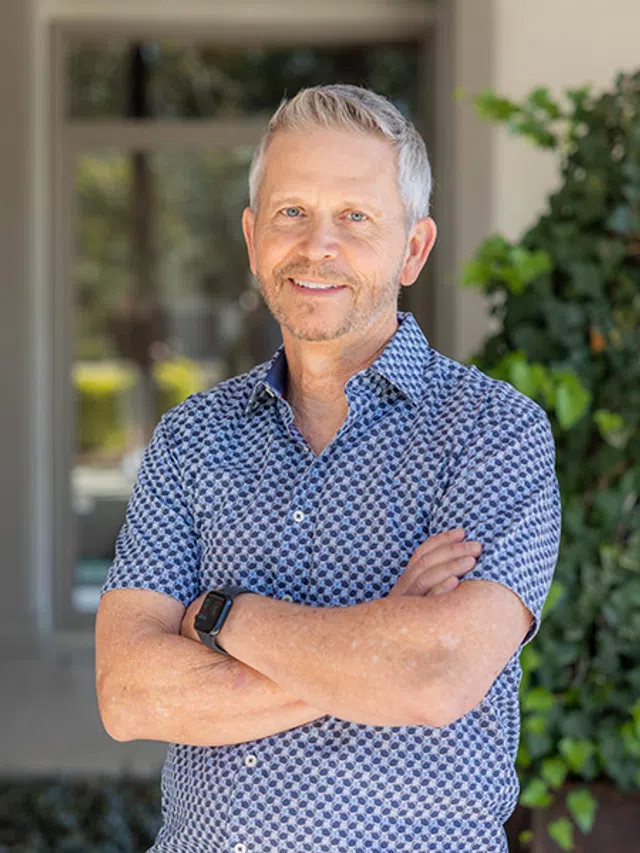 Smiling middle-aged man with gray hair and beard wearing patterned shirt and smartwatch, standing outdoors with arms crossed.