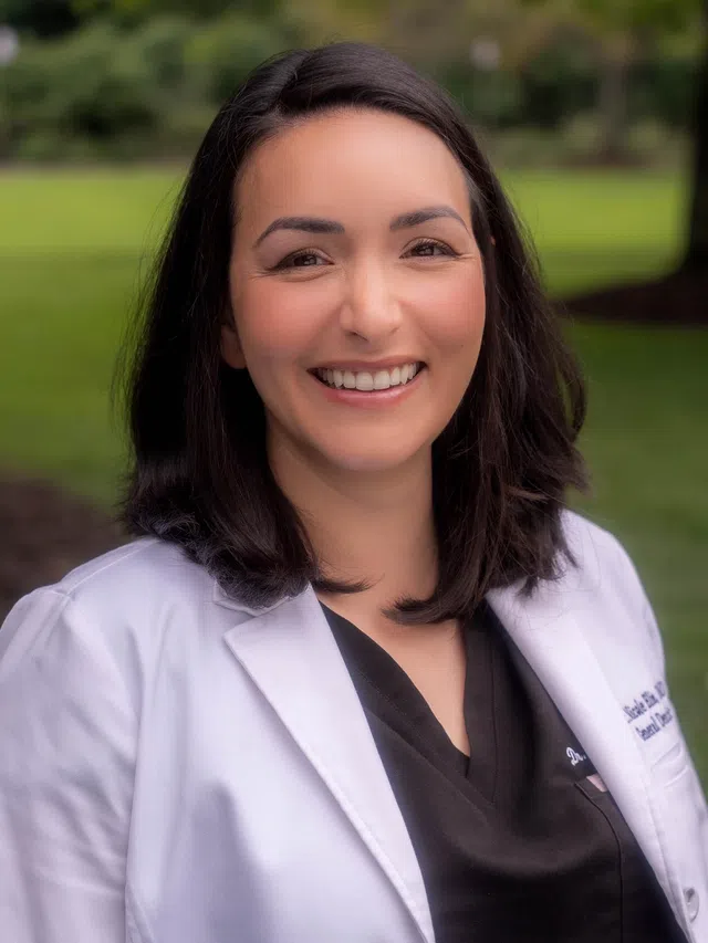 Smiling female dental medic in a white coat with name badge and dental association pins on a blue background