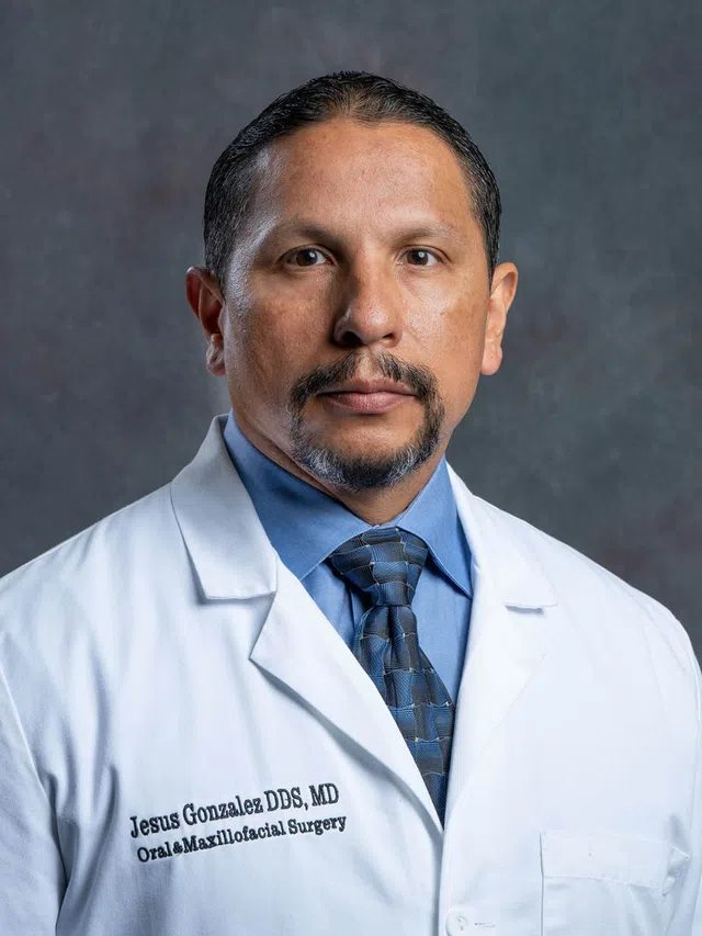 Portrait of a male doctor in a white coat and blue tie, with a focused expression against a gray backdrop.